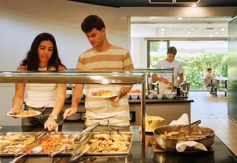 Students serving themselves at the buffet area of the Expanish Barcelona summer camp residence, with a variety of hot dishes available.