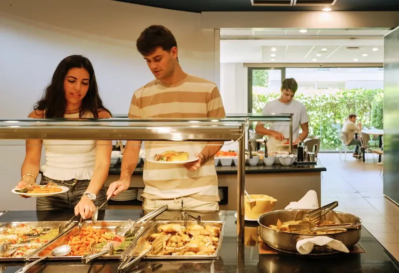 Students serving themselves at the buffet area of the Expanish Barcelona summer camp residence, with a variety of hot dishes available.