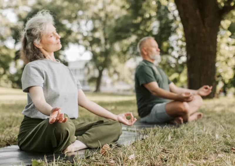 Two adults meditating outdoors on mats surrounded by greenery.