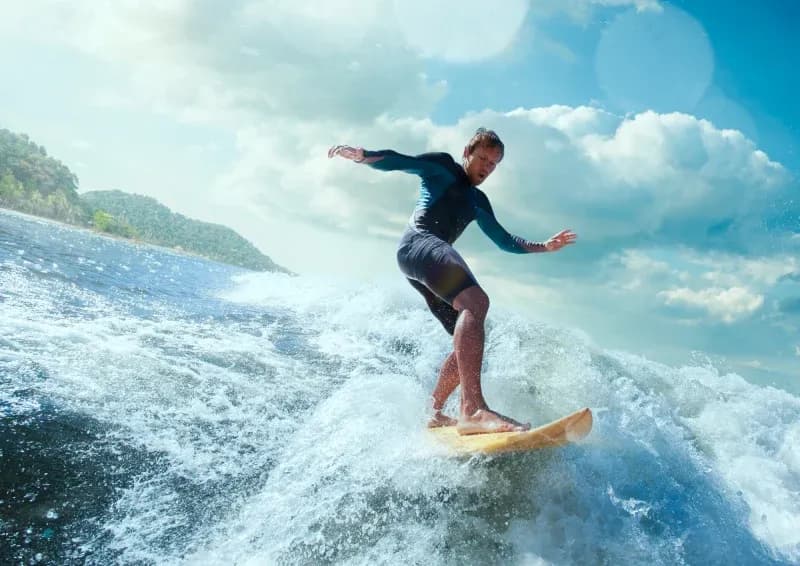 Student riding a wave on a surfboard with the ocean and coastline in the background.