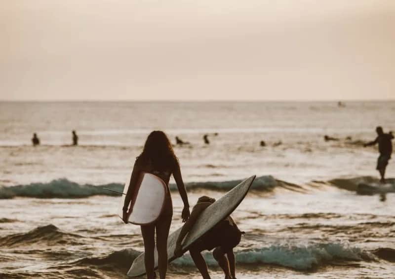 Young adults carrying surfboards into the ocean at sunset.