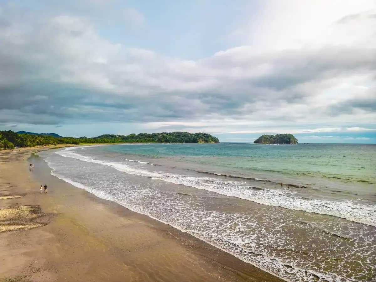 Wide view of a sandy beach in Samara with gentle waves, green hills, and small islands offshore.