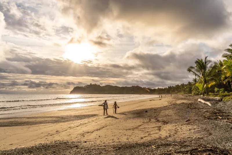 Wide view of a Costa Rica beach with palm trees, shoreline, and people walking at sunset.