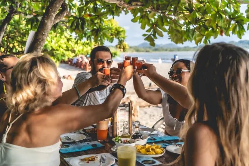 Group of students clinking drinks at a beachside table under tropical trees.