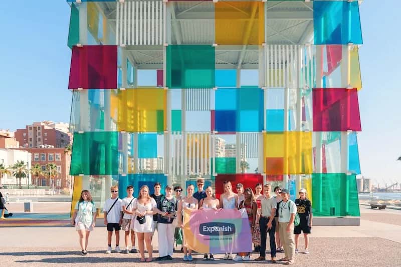 Expanish summer camp students posing with a flag in front of the colorful Centre Pompidou Málaga.